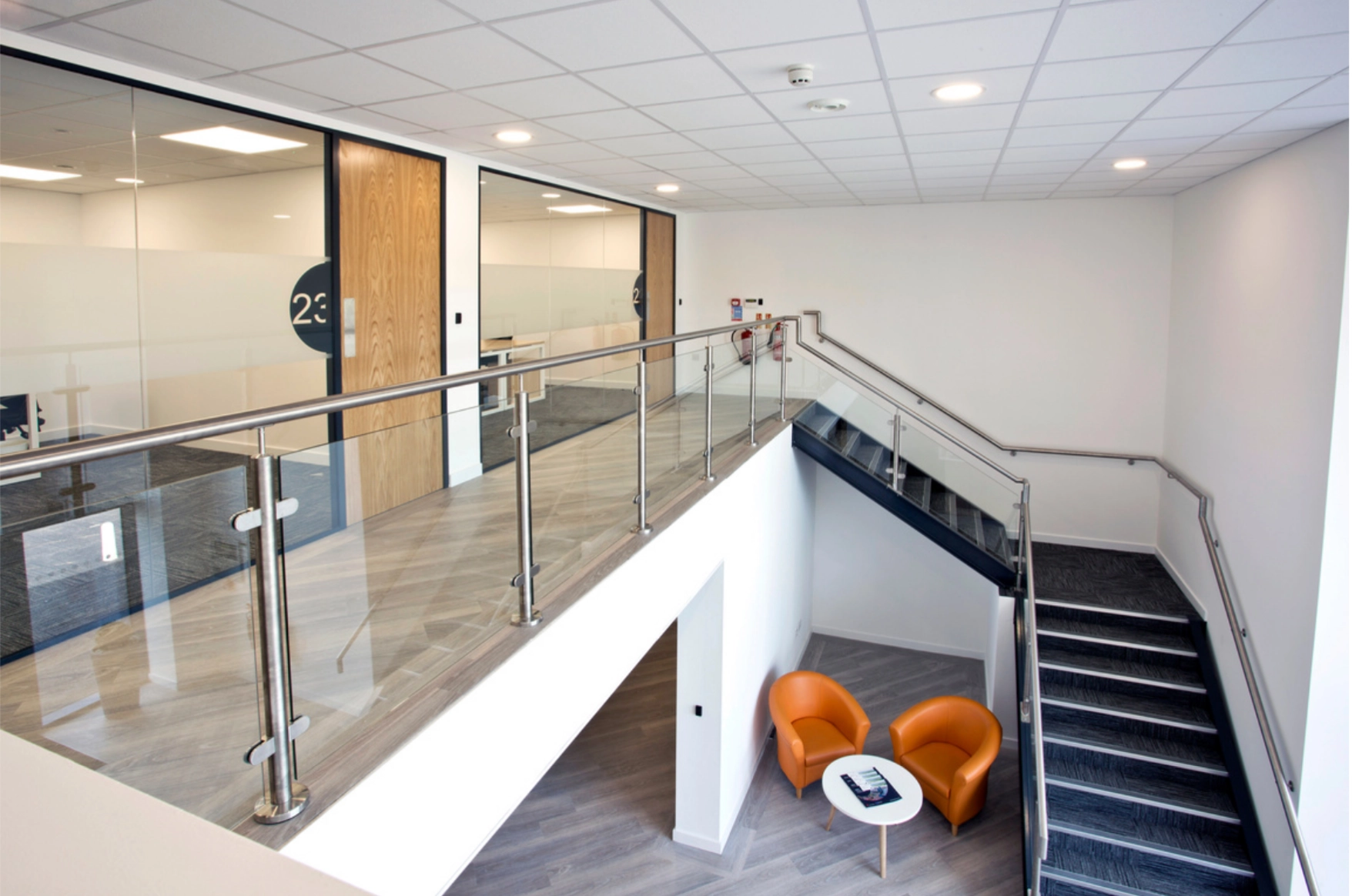 Modern office stairwell with glass and metal railing, two orange chairs and a small round table below.