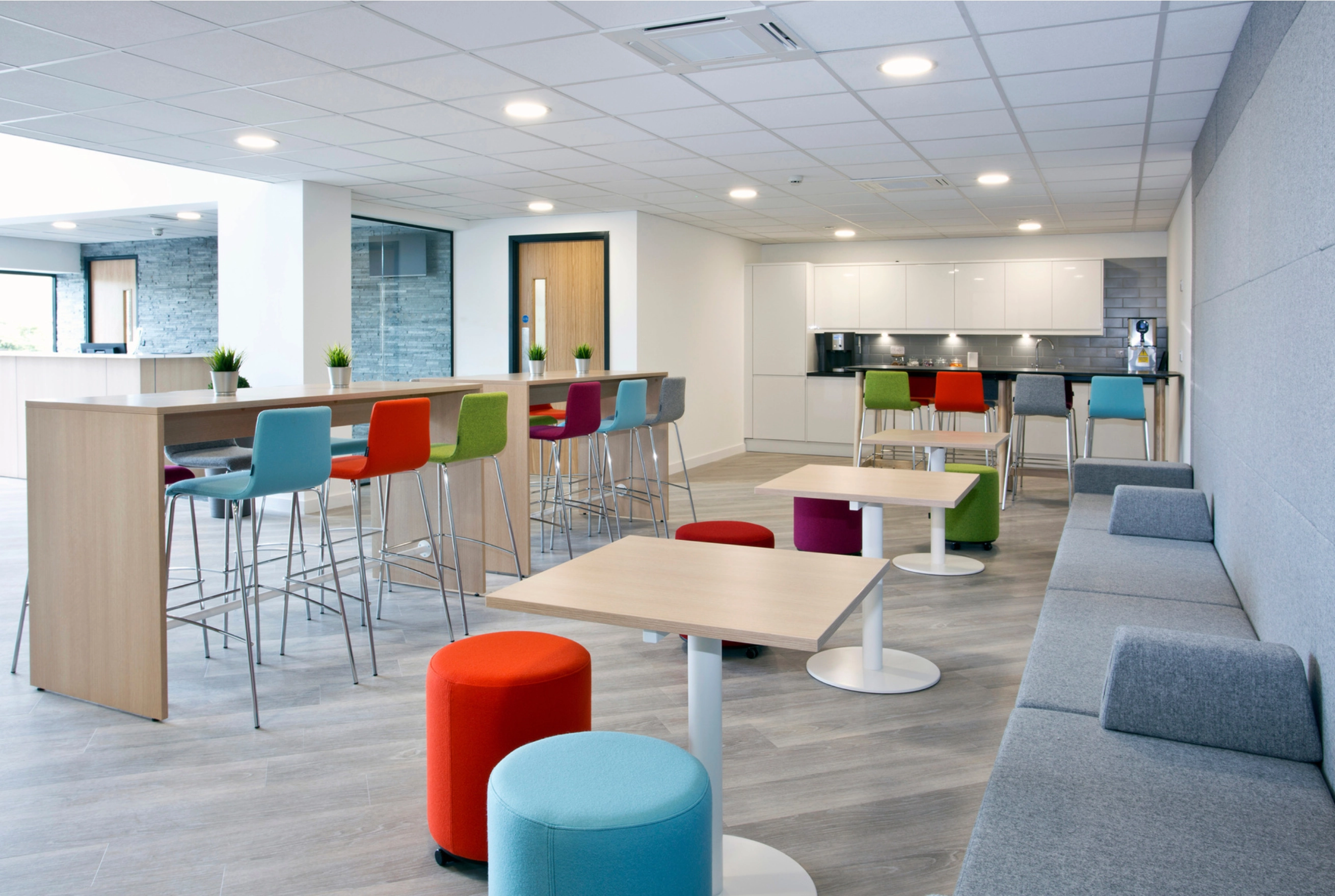 Modern office break room with colorful bar stools, gray sofa, small tables, and kitchenette with white cabinets.