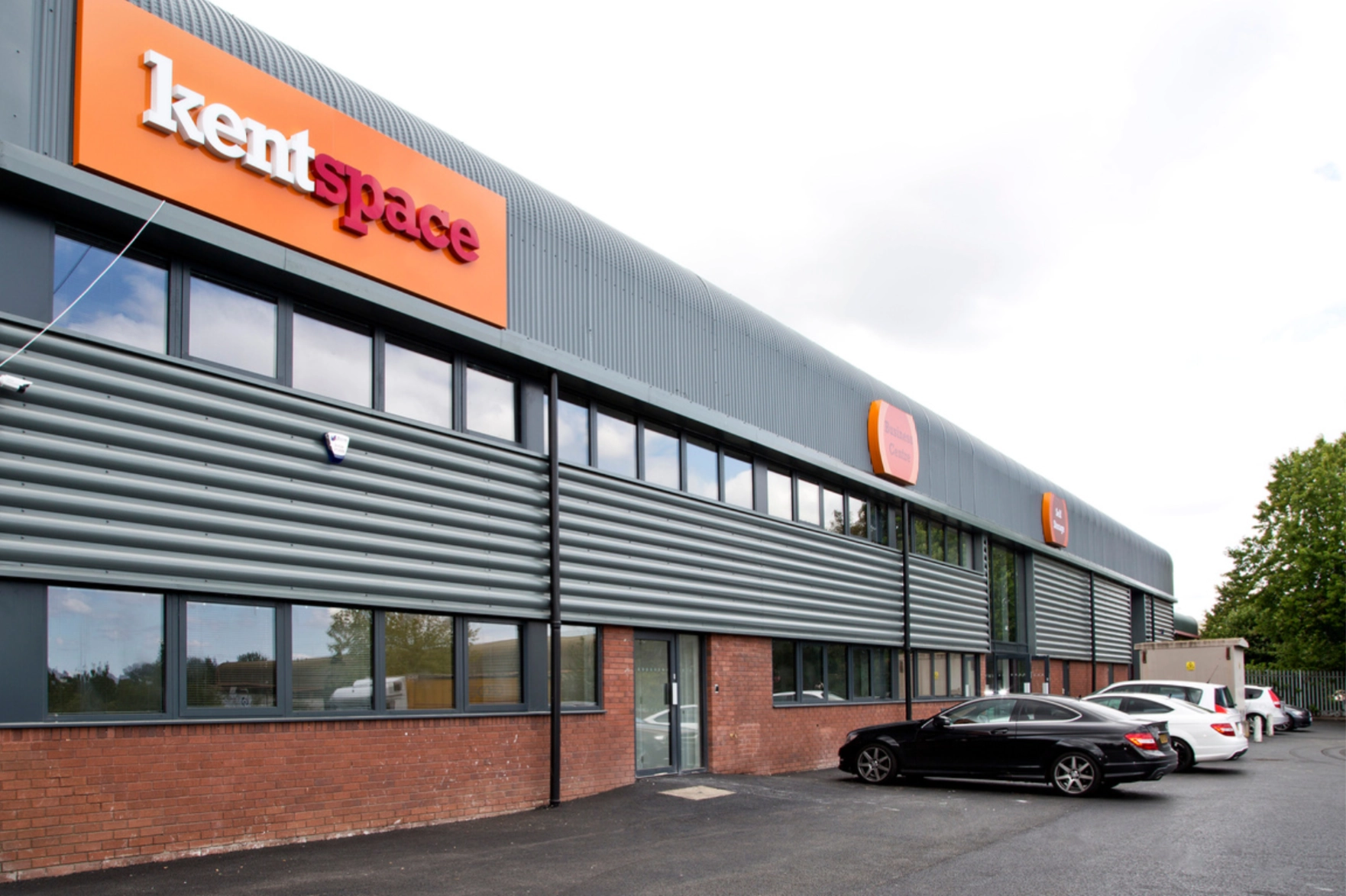 Exterior view of a modern industrial building with red brick lower walls, gray metal siding, large windows, and an orange sign reading 'kentspace', with cars parked in front.