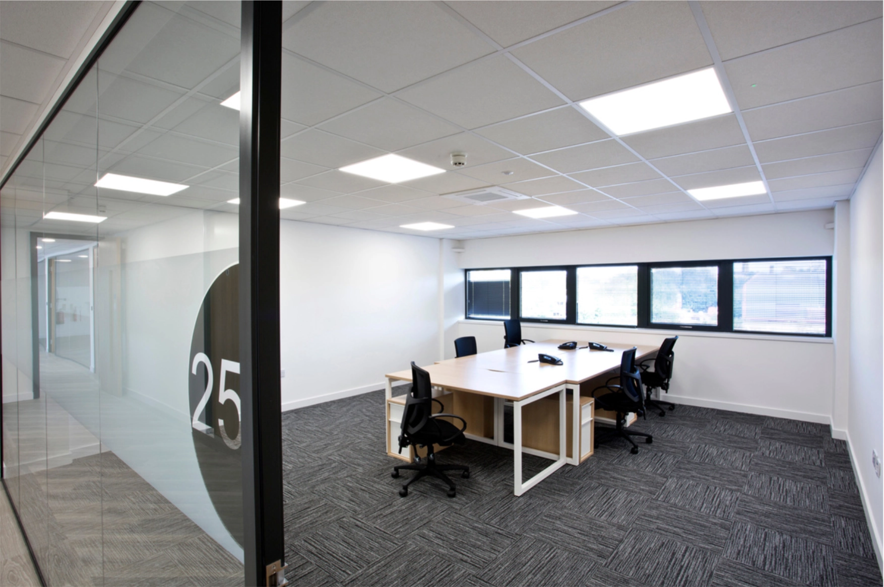 Modern office meeting room with four black chairs around two wooden tables, telephones on the tables, carpet floor, and large windows with blinds.