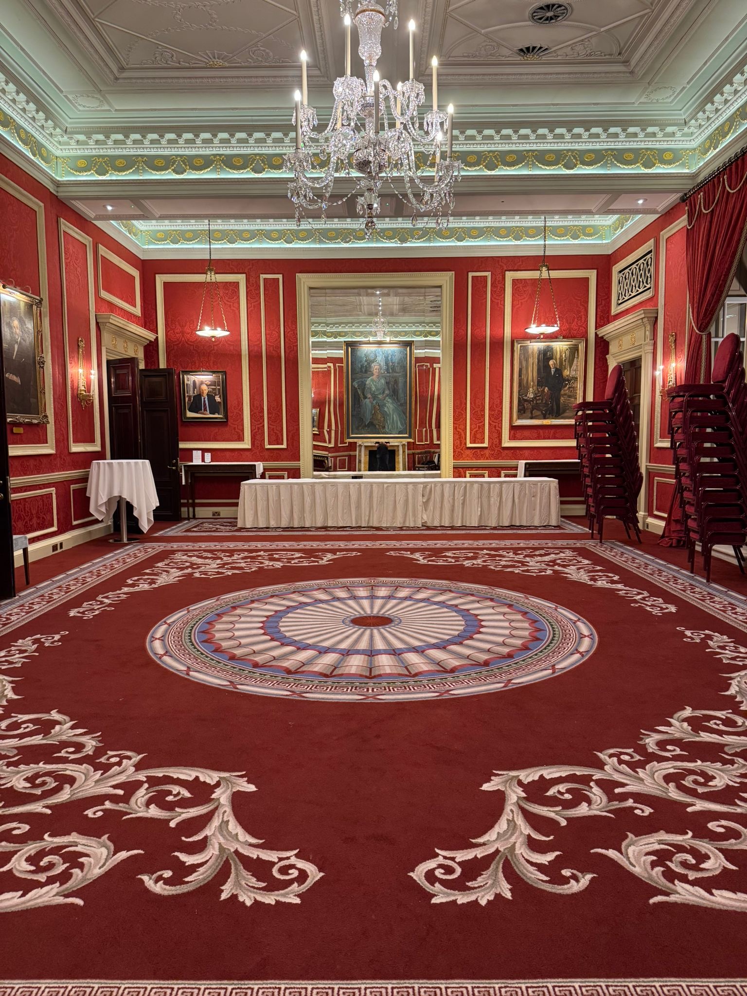 Elegant red and gold committee room with ornate carpet, large mirror, crystal chandelier, and stacked chairs.