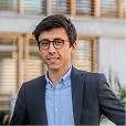 Homme souriant portant des lunettes et un costume bleu devant une façade moderne en béton.