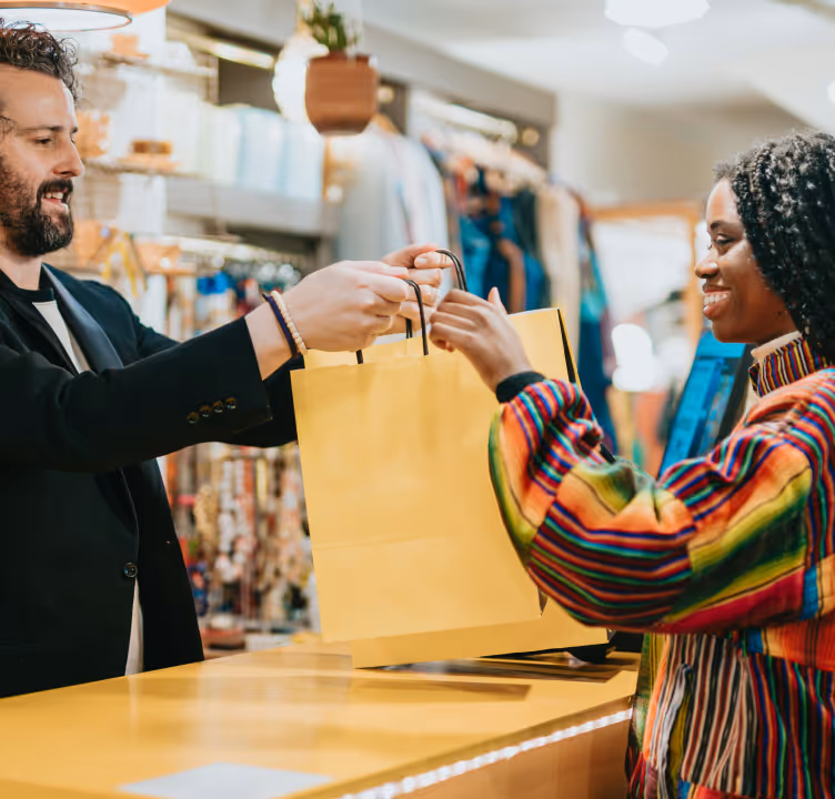 Un commerçant souriant remet un sac jaune à une cliente souriante portant un vêtement multicolore dans une boutique.