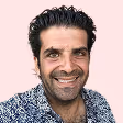 Portrait d'un homme souriant aux cheveux noirs bouclés, portant une chemise à motifs bleus et blancs, sur fond rose clair.