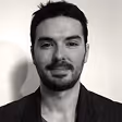 Portrait en noir et blanc d'un homme avec une barbe légère et des cheveux courts, portant une veste sombre.