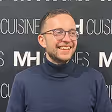 Un homme souriant portant des lunettes et un pull à col roulé bleu marine devant un arrière-plan avec du texte.