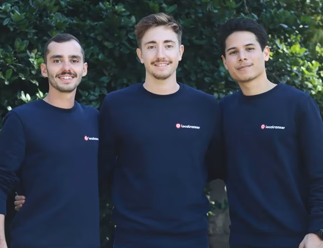 Trois jeunes hommes souriants portant des pulls bleu marine avec le logo Localranker, posant devant un feuillage vert.