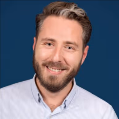 Portrait d'un homme souriant avec une barbe et une chemise rayée claire sur fond bleu.