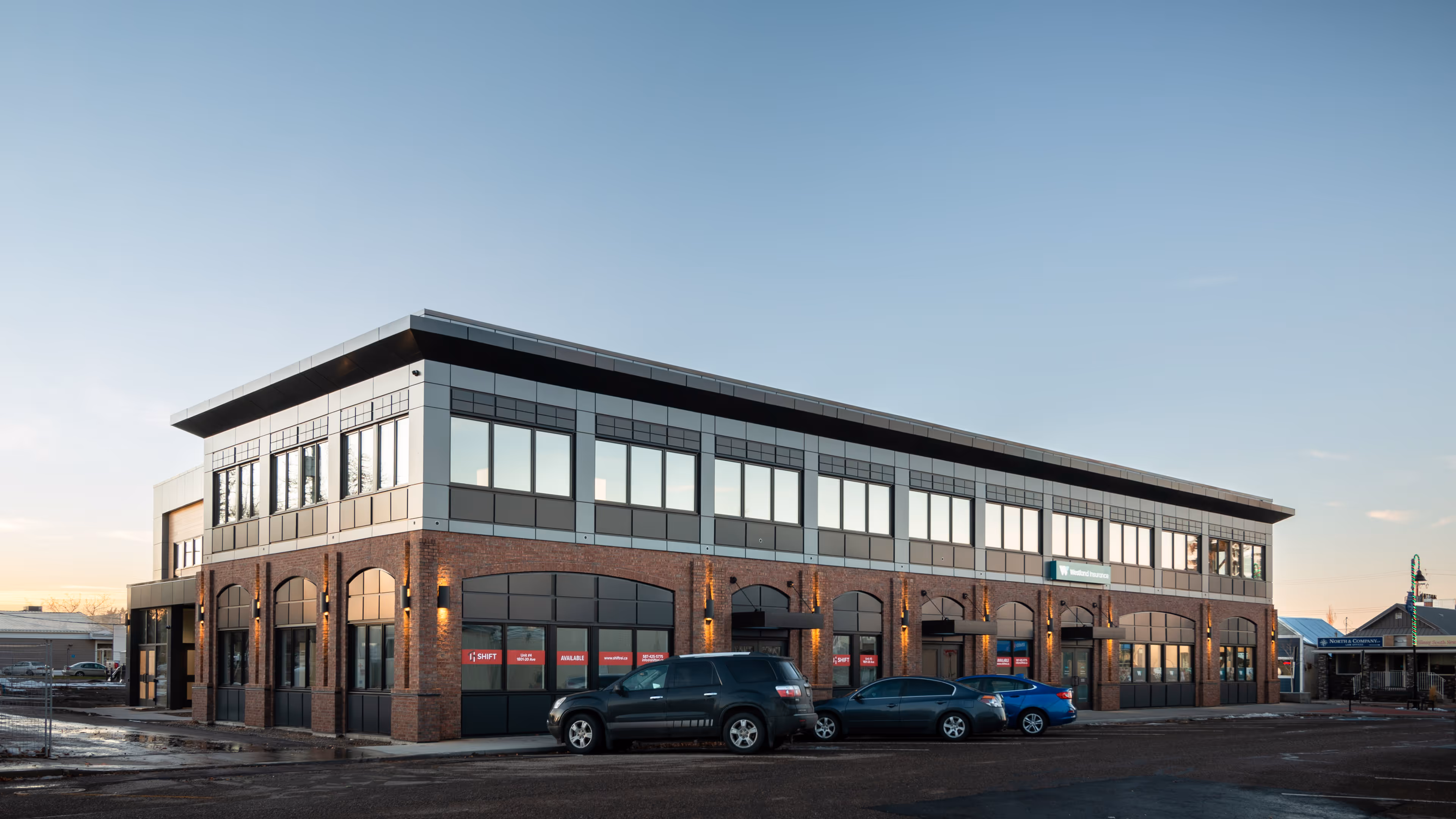 Two-story commercial building with large windows and brick facade at dusk with three parked cars in front.