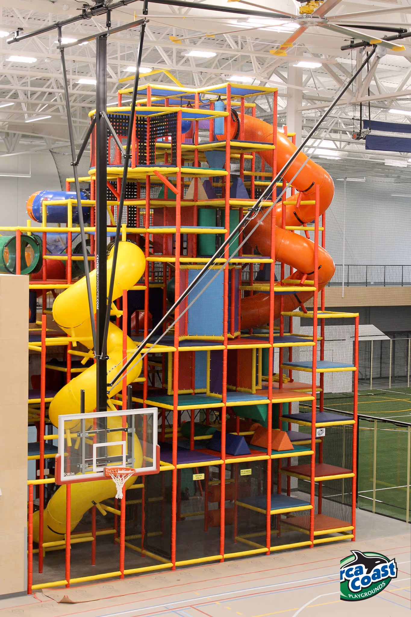Indoor multi-level playground structure with yellow and orange spiral slides and colorful climbing nets in a sports court.