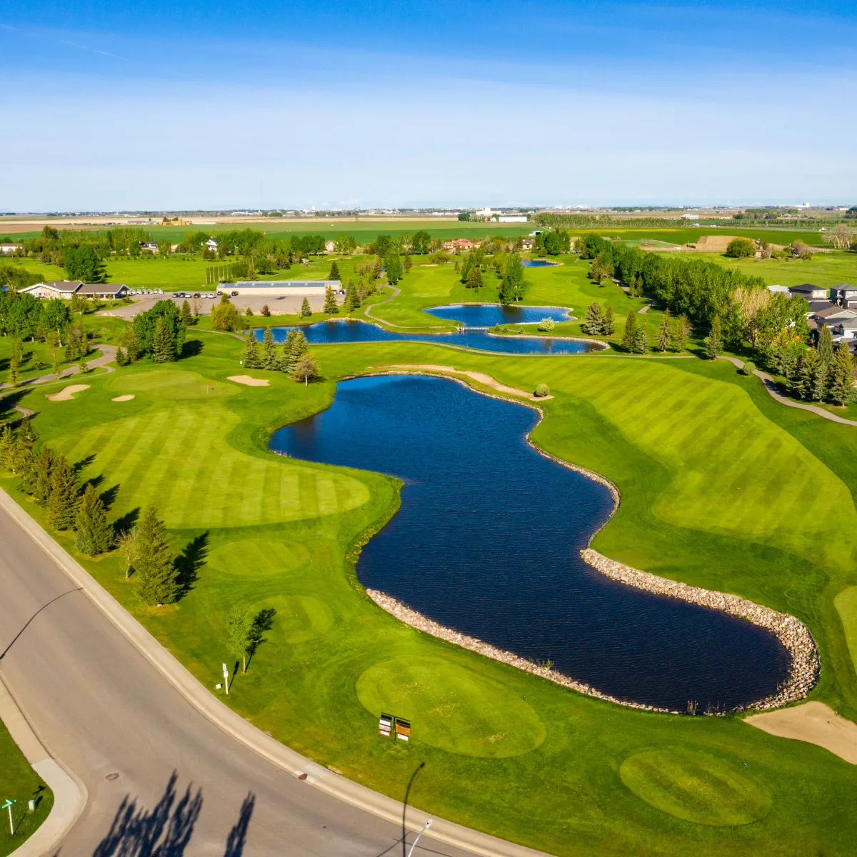 Aerial view of a golf course featuring green fairways, multiple water hazards, sand bunkers, and surrounding trees under a clear blue sky.