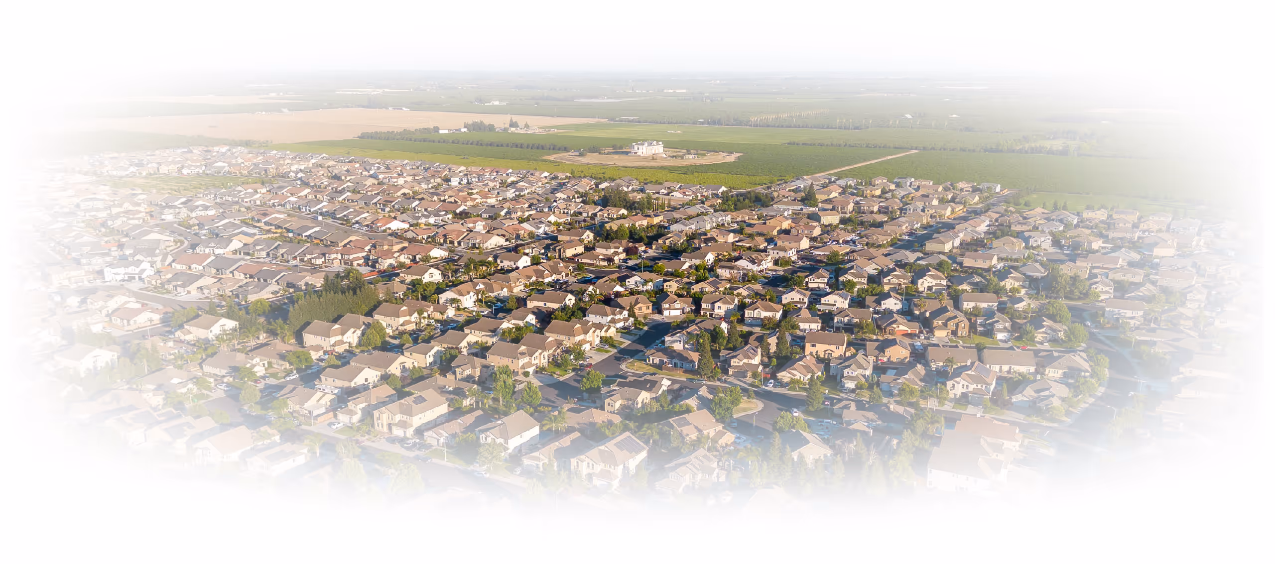 Aerial view of a suburban neighborhood with numerous houses, tree-lined streets, and farmland in the background.