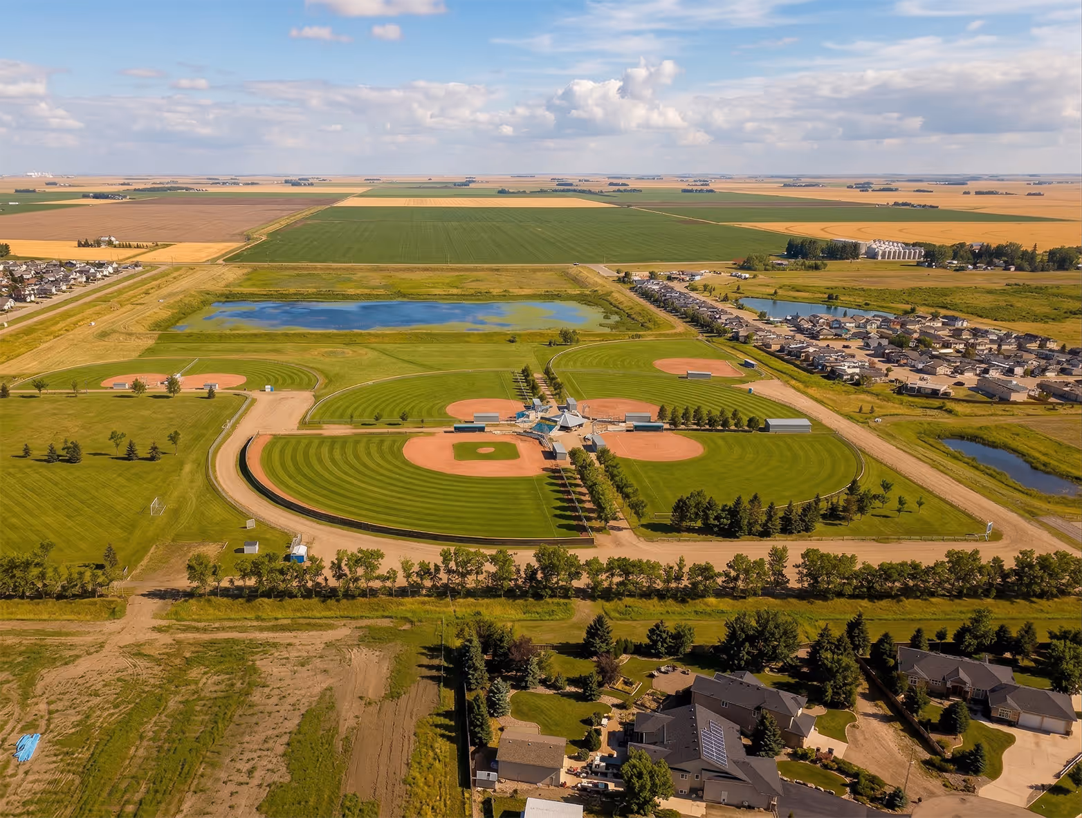 Aerial view of a baseball complex with multiple fields, surrounded by farmland and residential areas under a partly cloudy sky.