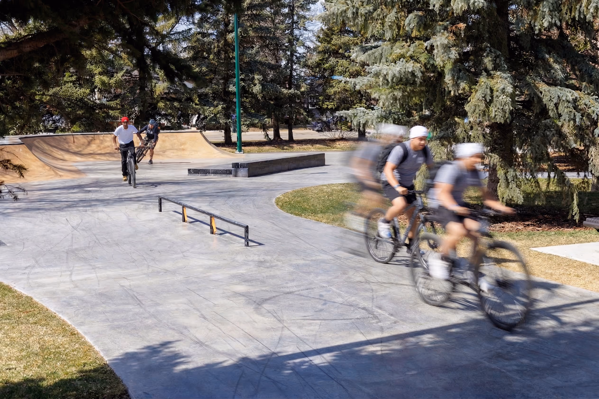 Two cyclists riding in a skate park with ramps and railings amidst trees, one cyclist blurred in motion.