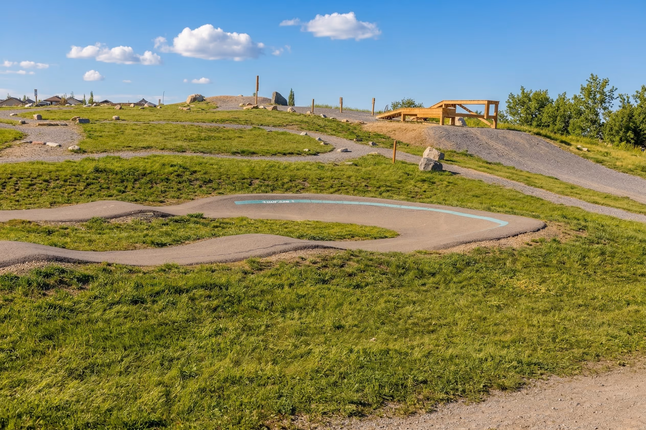 Outdoor bike park with paved winding track, green grass, gravel paths, wooden ramp, and a clear blue sky with clouds.