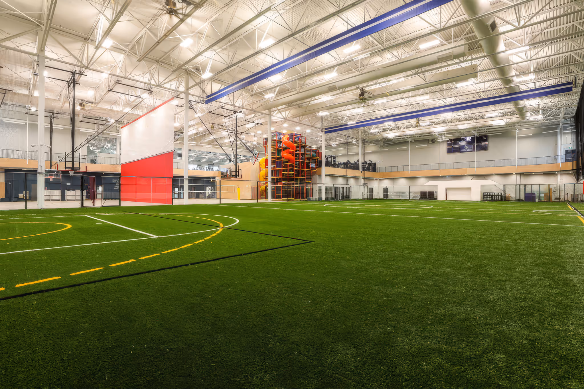 Indoor sports facility with artificial turf field marked for soccer and basketball, featuring a climbing structure with slides.