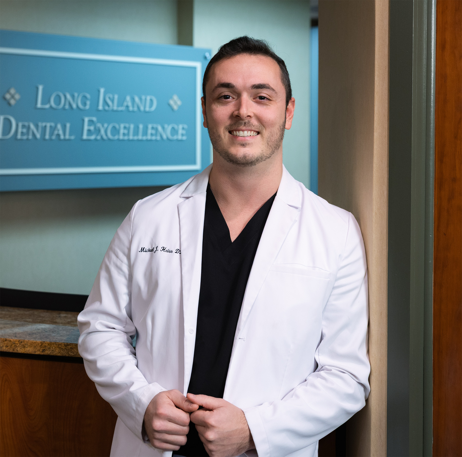 Smiling male dentist in a white coat standing in front of a sign that reads Long Island Dental Excellence.