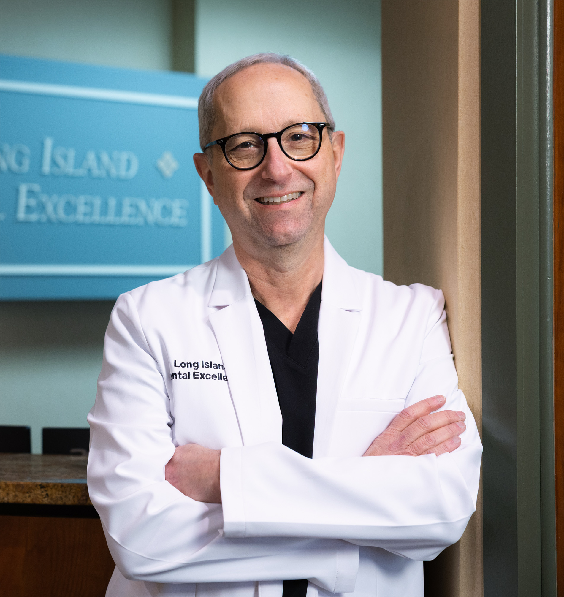 Smiling middle-aged man wearing glasses and a white medical coat with 'Long Island Dental Excellence' embroidered, standing with arms crossed.