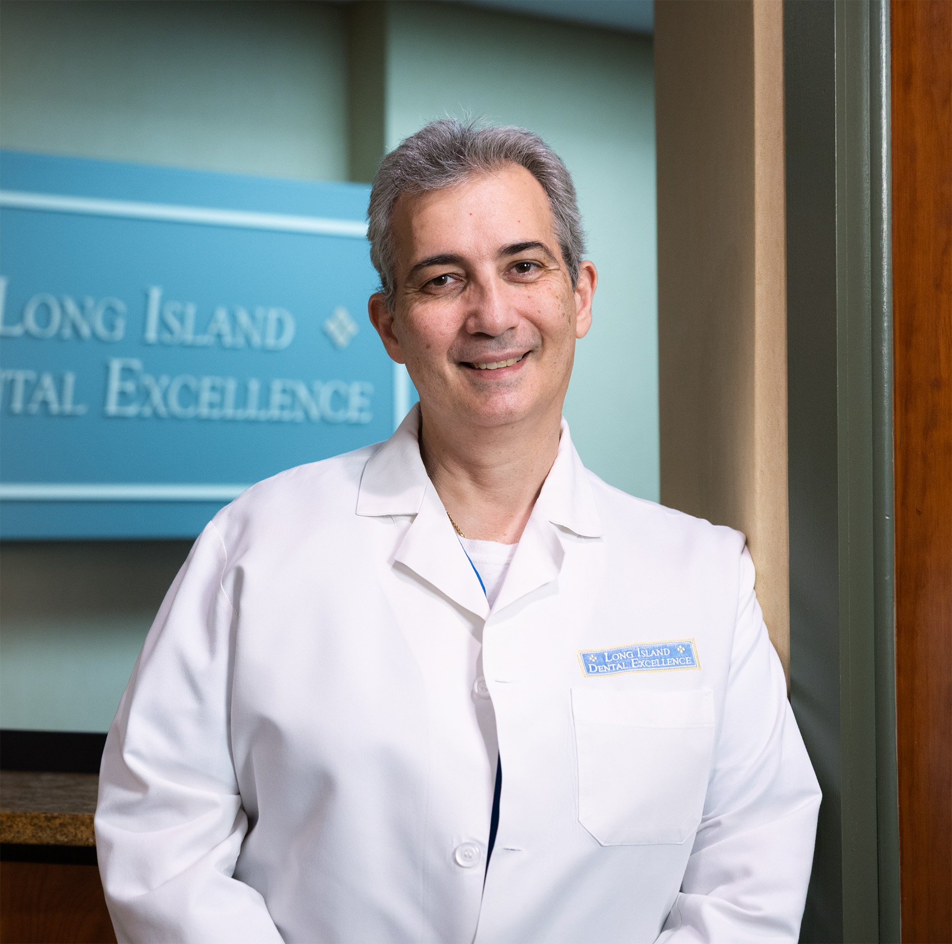 Smiling male dentist standing indoors wearing a white coat with a Long Island Dental Excellence badge.