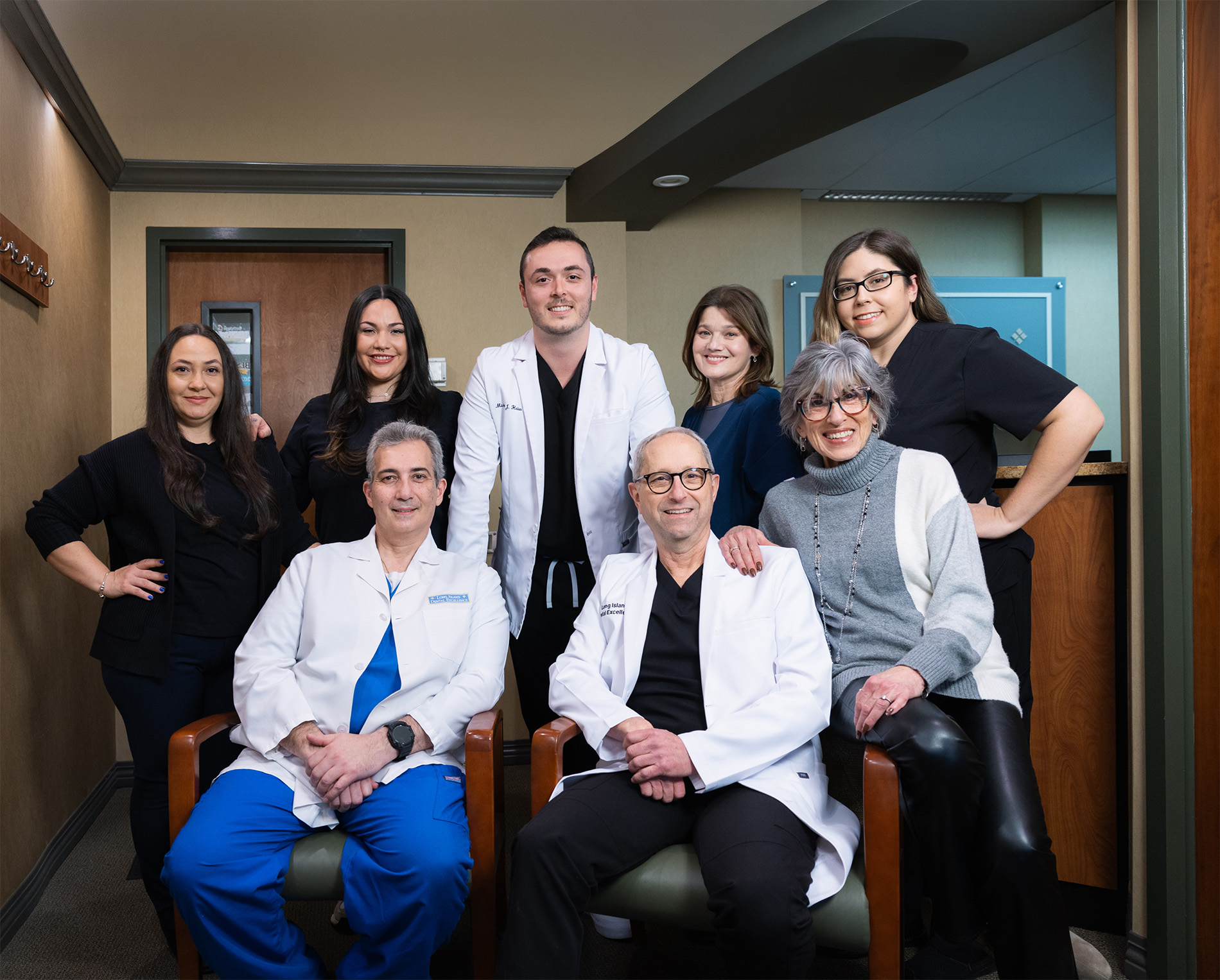 Group portrait of eight medical professionals in a clinic waiting room, two seated men in white coats and six standing people smiling.