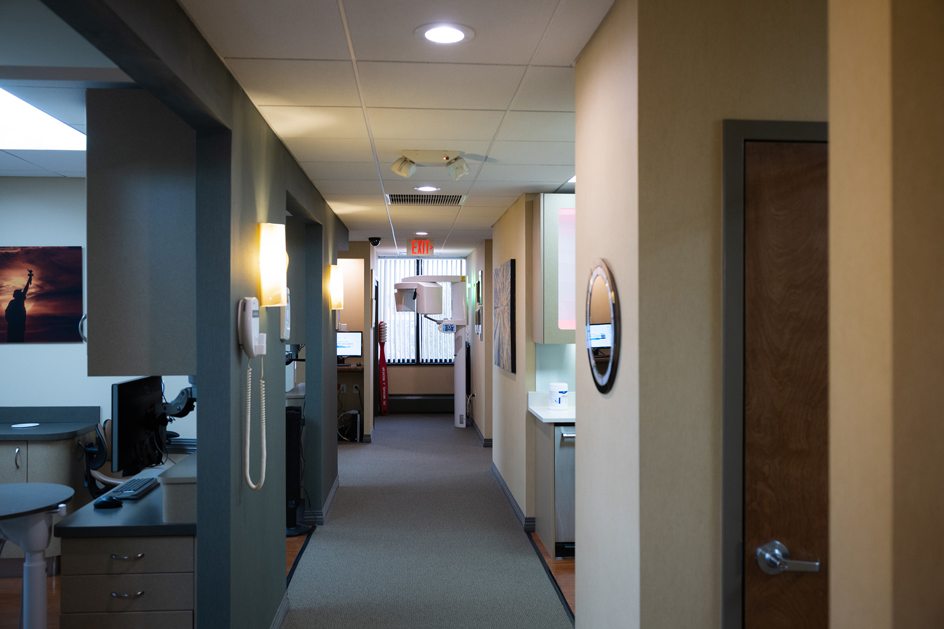 Hallway in a modern dental or medical office with beige walls, computer workstations, and an exit sign at the end.