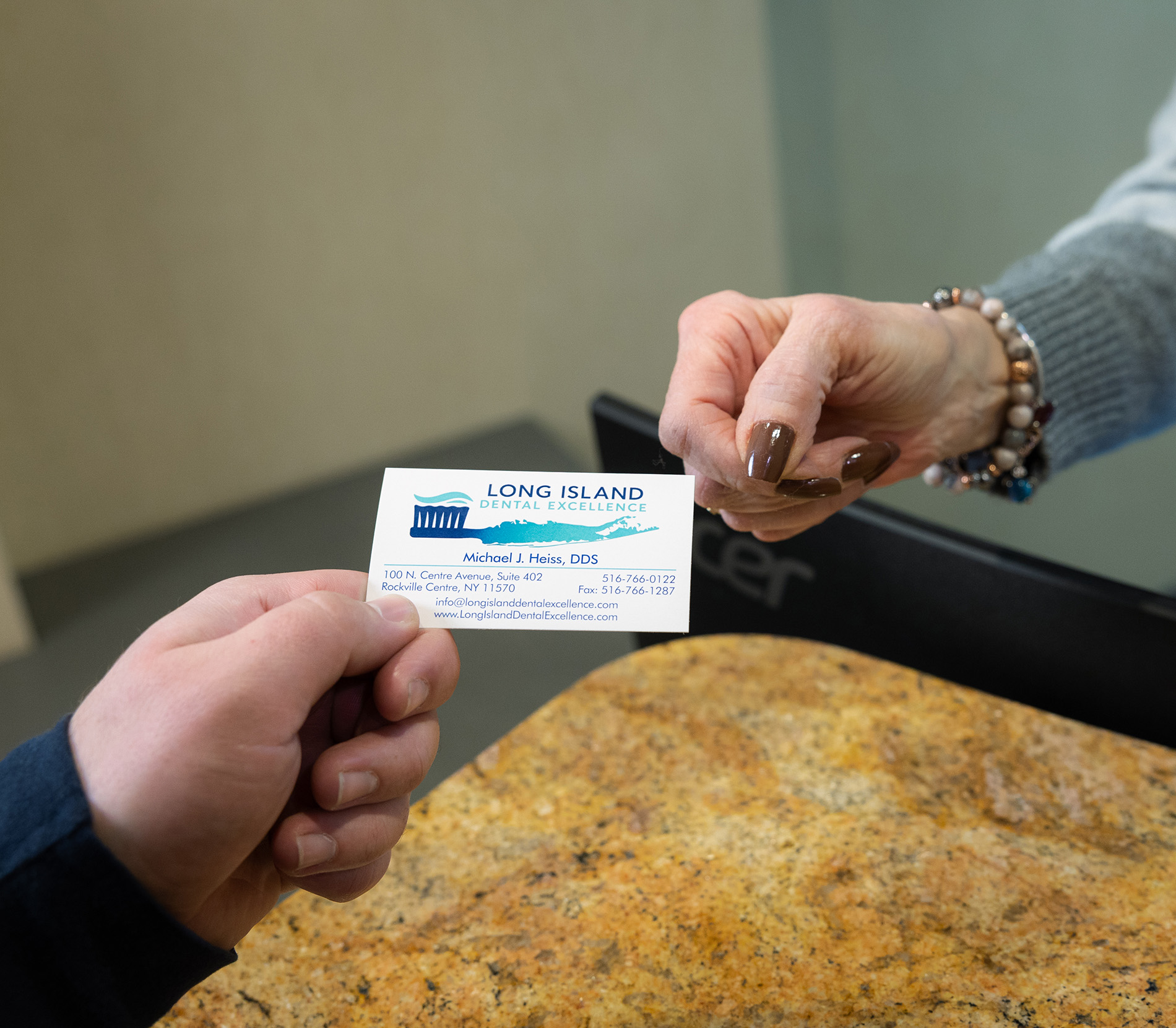 Two hands exchanging a business card for Long Island Dental Excellence with contact details.