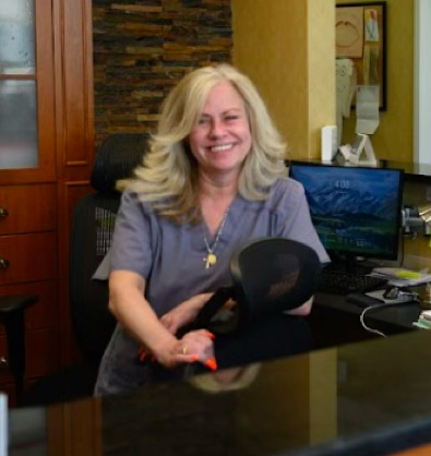 Smiling woman with blonde hair wearing scrubs, sitting at a desk with a computer in the background.