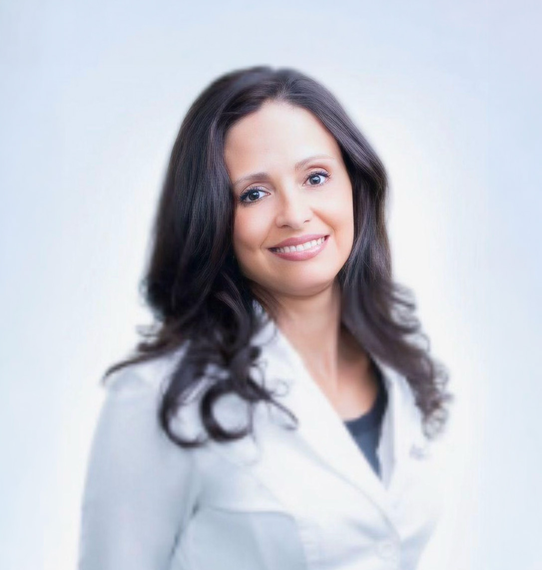 Smiling woman with long dark hair wearing a white blazer over a black top against a light background.