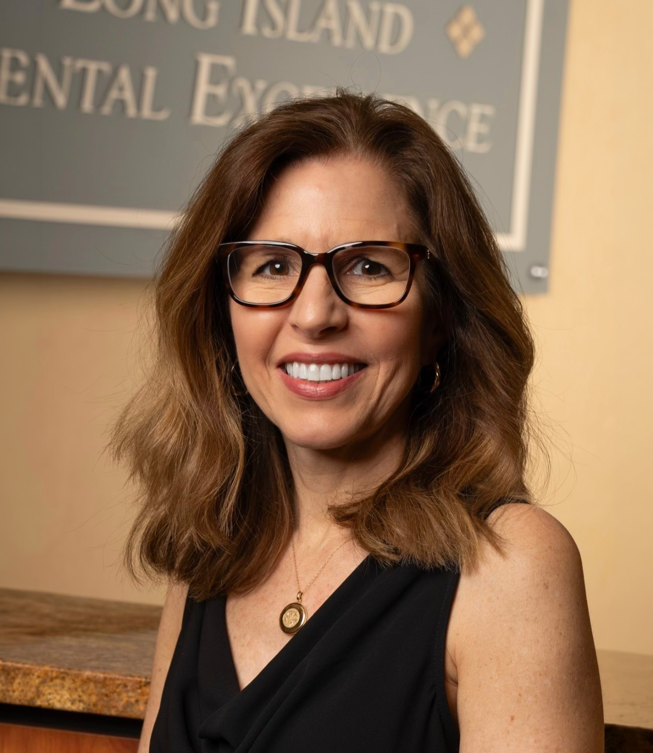 Smiling woman with glasses and shoulder-length brown hair in a black sleeveless top, standing indoors.