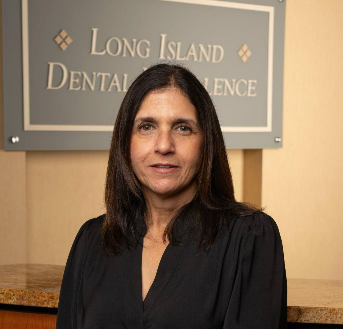 Woman with dark hair wearing a black top standing in front of a Long Island Dental Excellence sign.