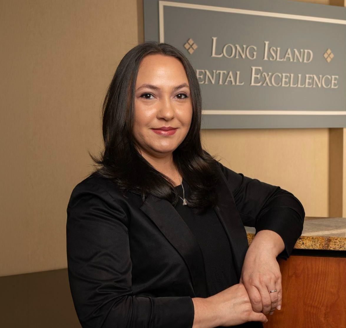 Woman with dark hair wearing a black blazer, leaning on a counter in front of a sign that reads Long Island Dental Excellence.