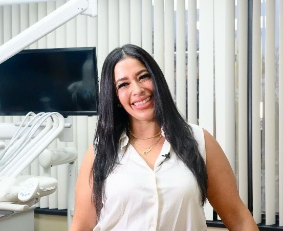 Smiling woman with long dark hair wearing a white blouse standing in a dental office with equipment and a monitor in the background.