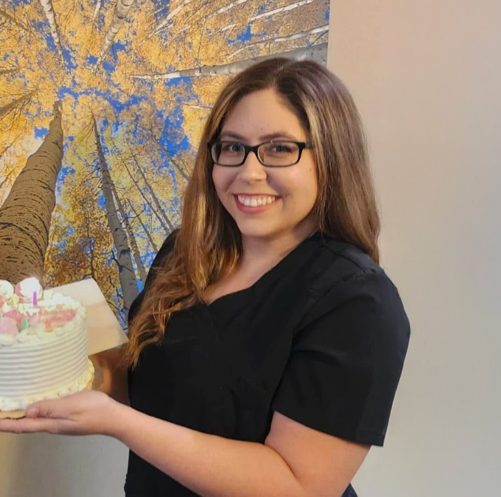 Smiling woman with glasses holding a decorated birthday cake with a lit candle, standing indoors in front of a wall mural of tall trees with yellow leaves and a blue sky.