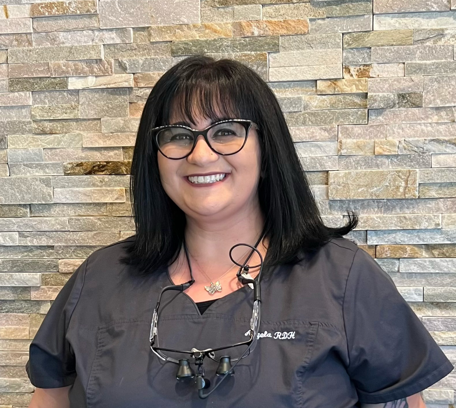 Smiling woman with black hair, glasses, and dental loupes around her neck wearing a dark scrub top in front of a stone wall.