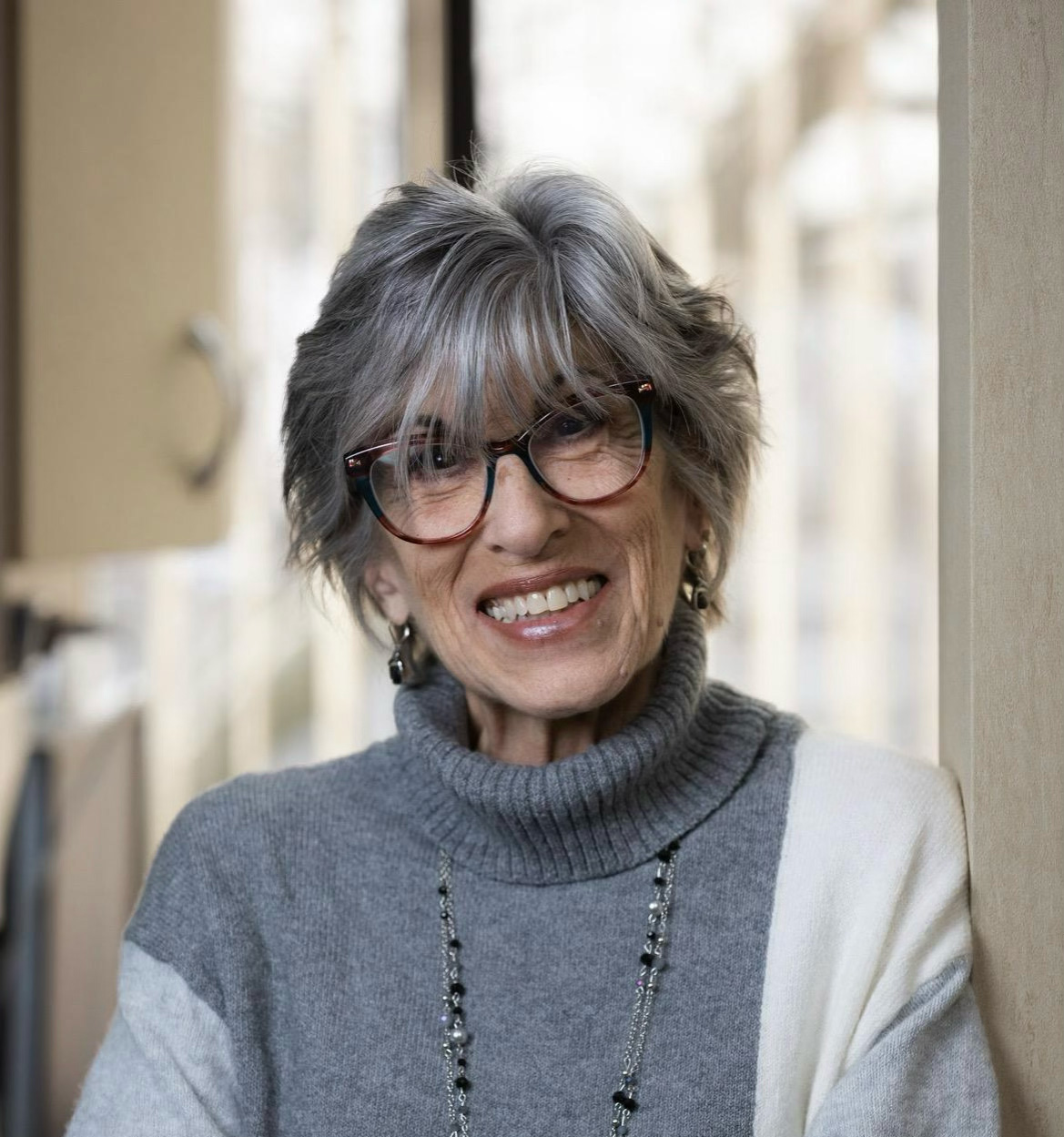 Smiling older woman with gray hair, wearing glasses, a gray and white sweater, and a beaded necklace.