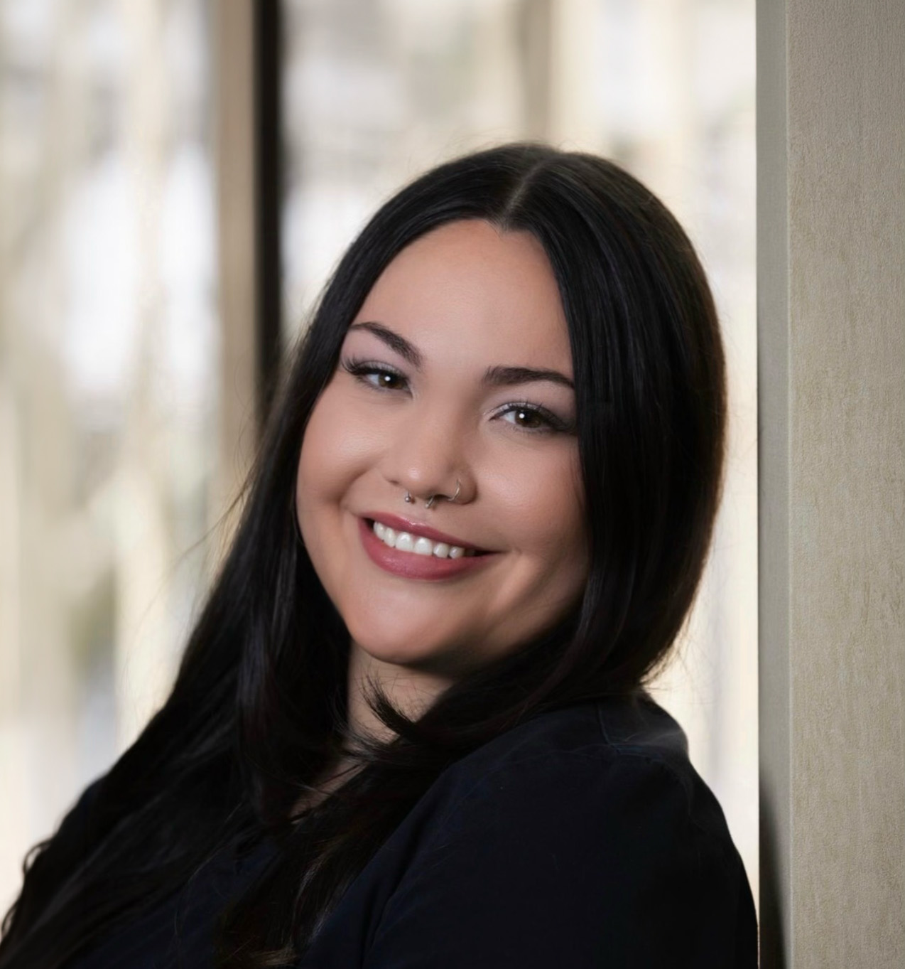 Smiling woman with long black hair leaning against a light-colored wall, wearing a black top and septum nose rings.