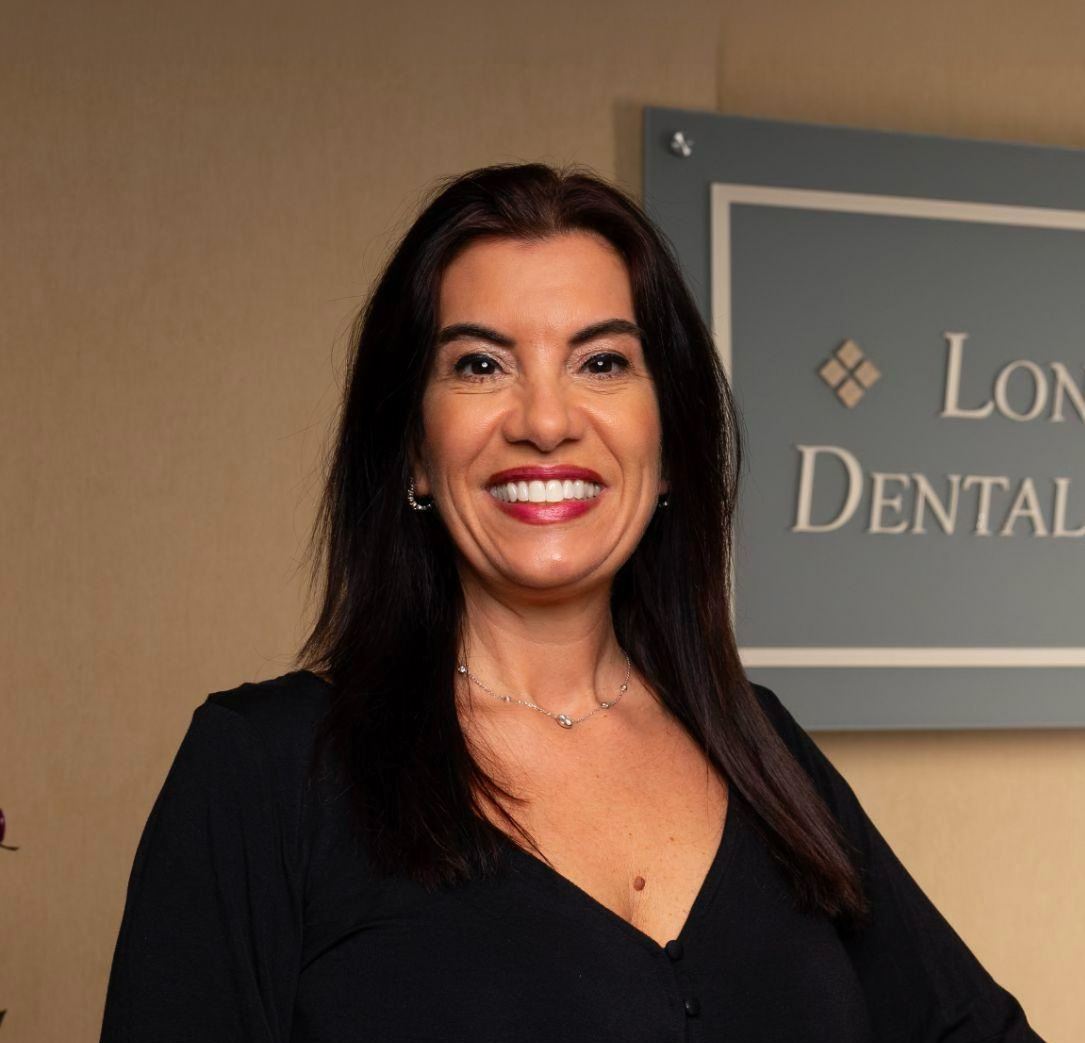 Smiling woman with dark hair wearing a black top standing in front of a dental clinic sign.
