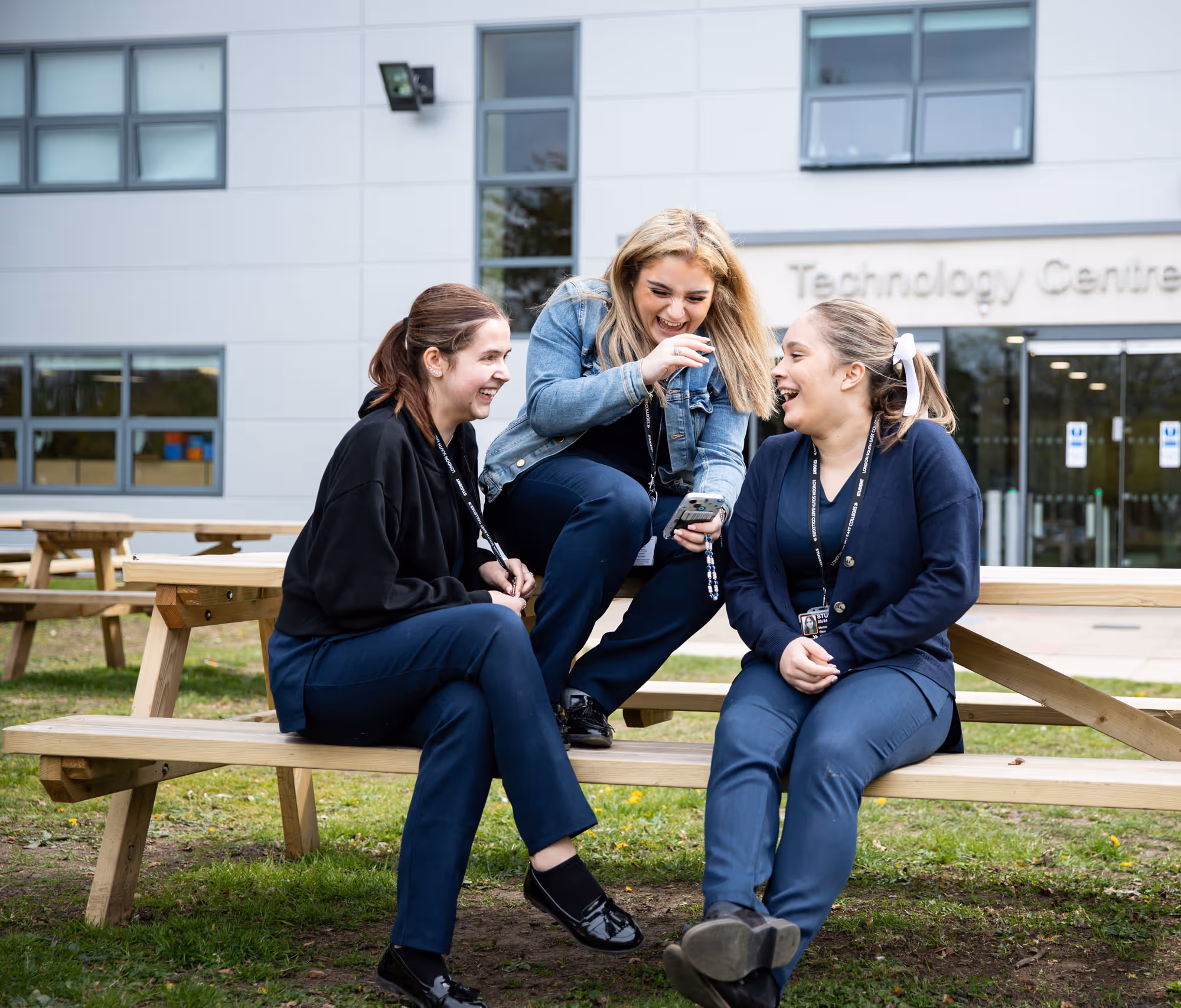 Three young women sitting and laughing on a wooden picnic bench outside a building labeled Technology Centre.