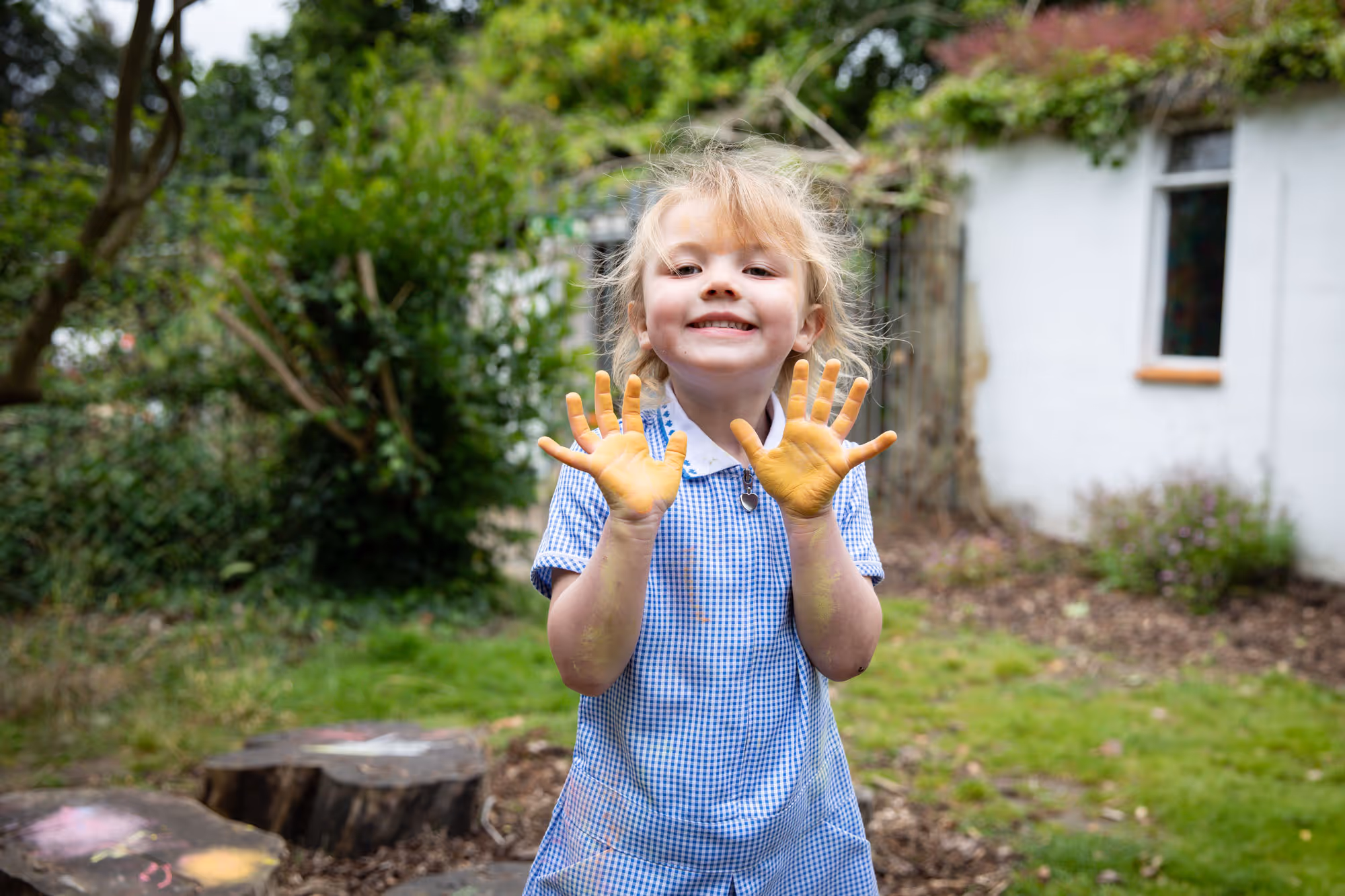Smiling young girl in a blue checkered dress showing her hands covered in yellow paint outdoors.