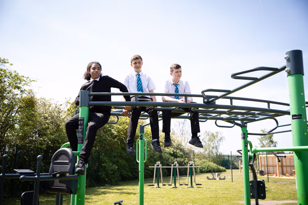 Three schoolboys in uniform sitting and leaning on green playground climbing equipment outdoors on a sunny day.