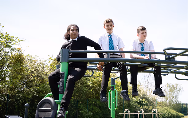 Three teenagers in school uniforms sitting and leaning on green playground monkey bars outdoors.