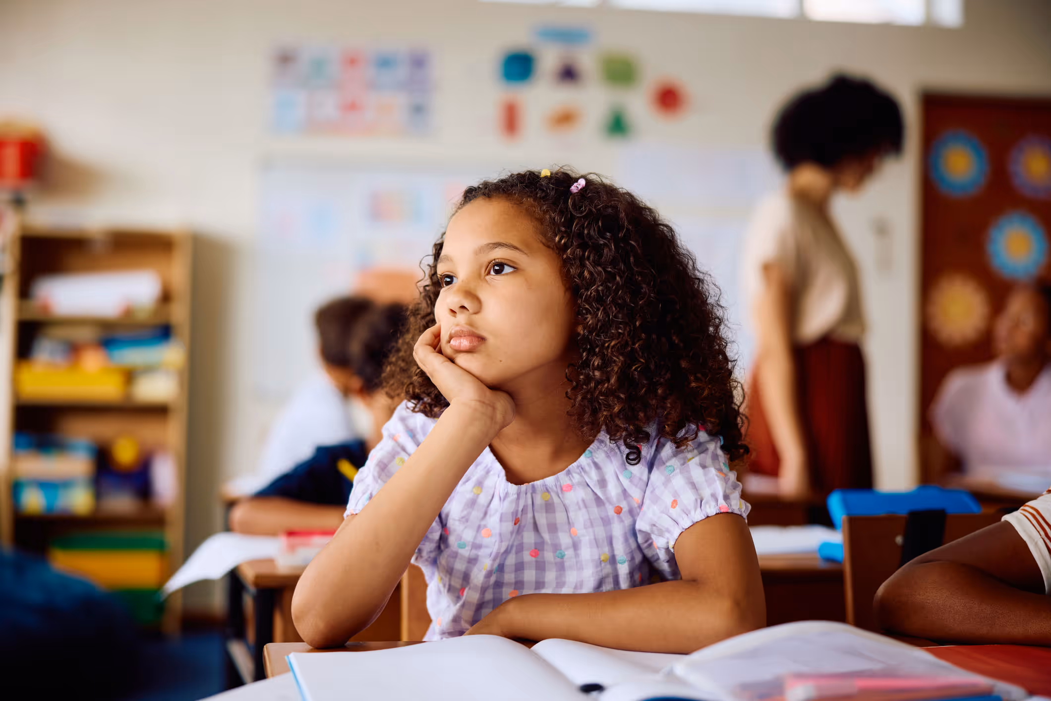 Young girl with curly hair resting her chin on hand, sitting at a desk in a classroom.