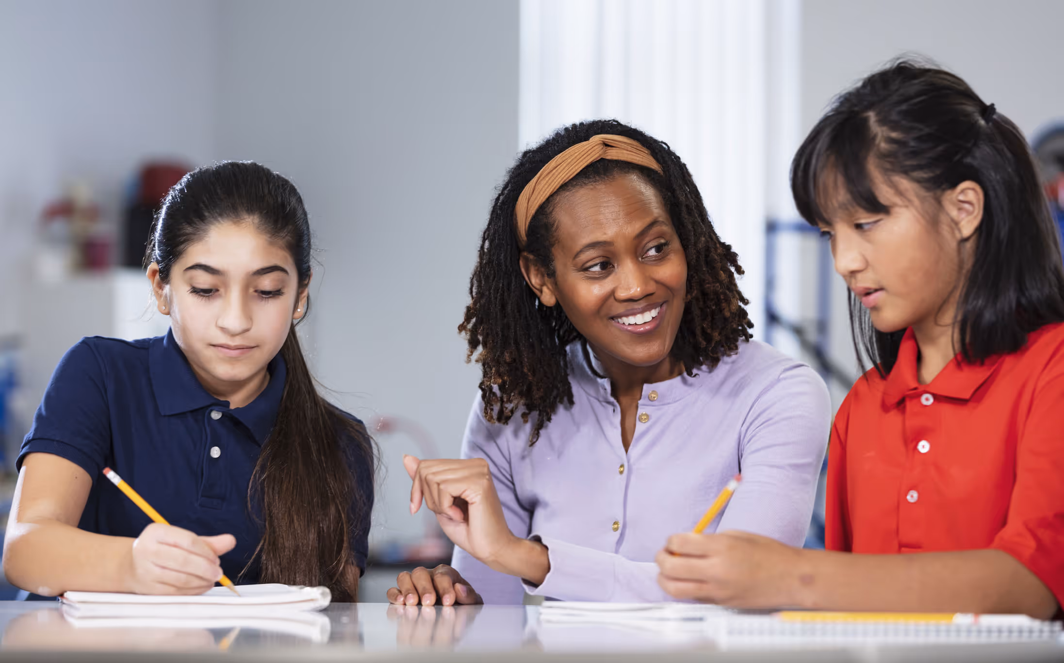 Teacher smiling and assisting two girls working with pencils and notebooks at a table.
