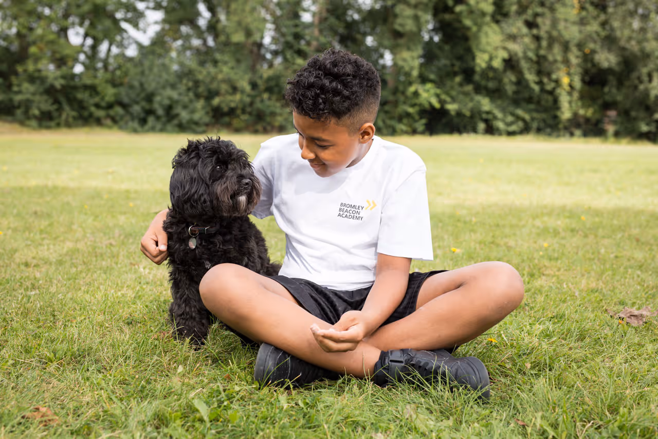 Boy in a white Bromley Beacon Academy T-shirt sitting cross-legged on grass with a black dog, outdoors.