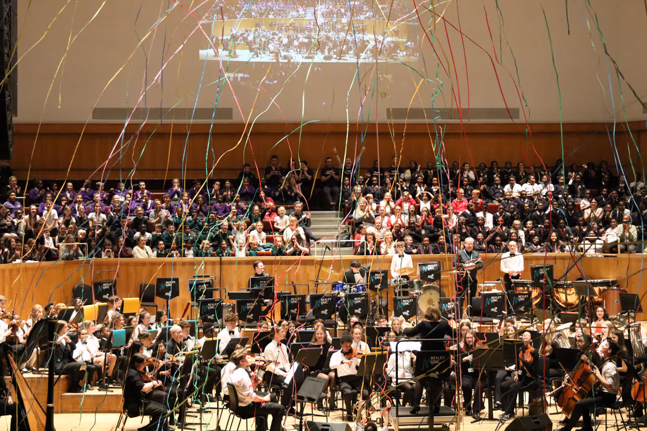 Orchestra performing in front of a large audience with colorful streamers falling from above in a concert hall.