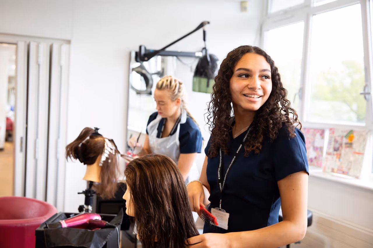 Two young women practicing hairstyling in a bright salon, one combing a mannequin's wet hair and the other applying hair color in the background.