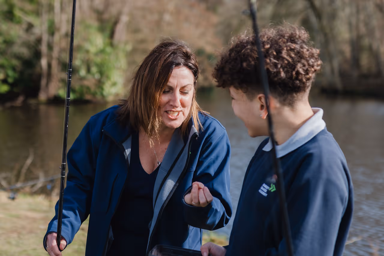Woman and young person smiling and holding fishing rods near a riverbank on a sunny day.