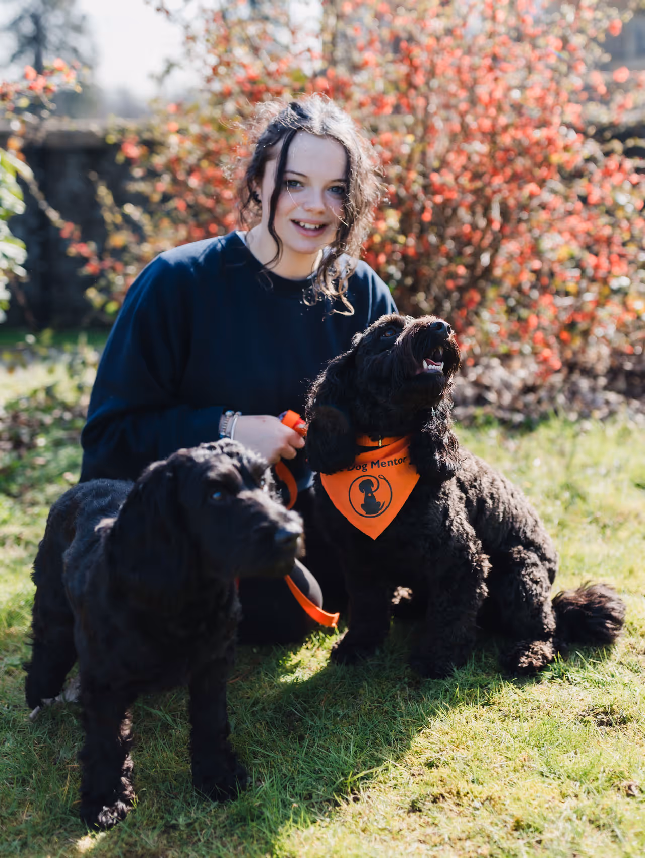 Smiling young woman outdoors with two black therapy dogs, one wearing an orange 'Dog Mentor' bandana.