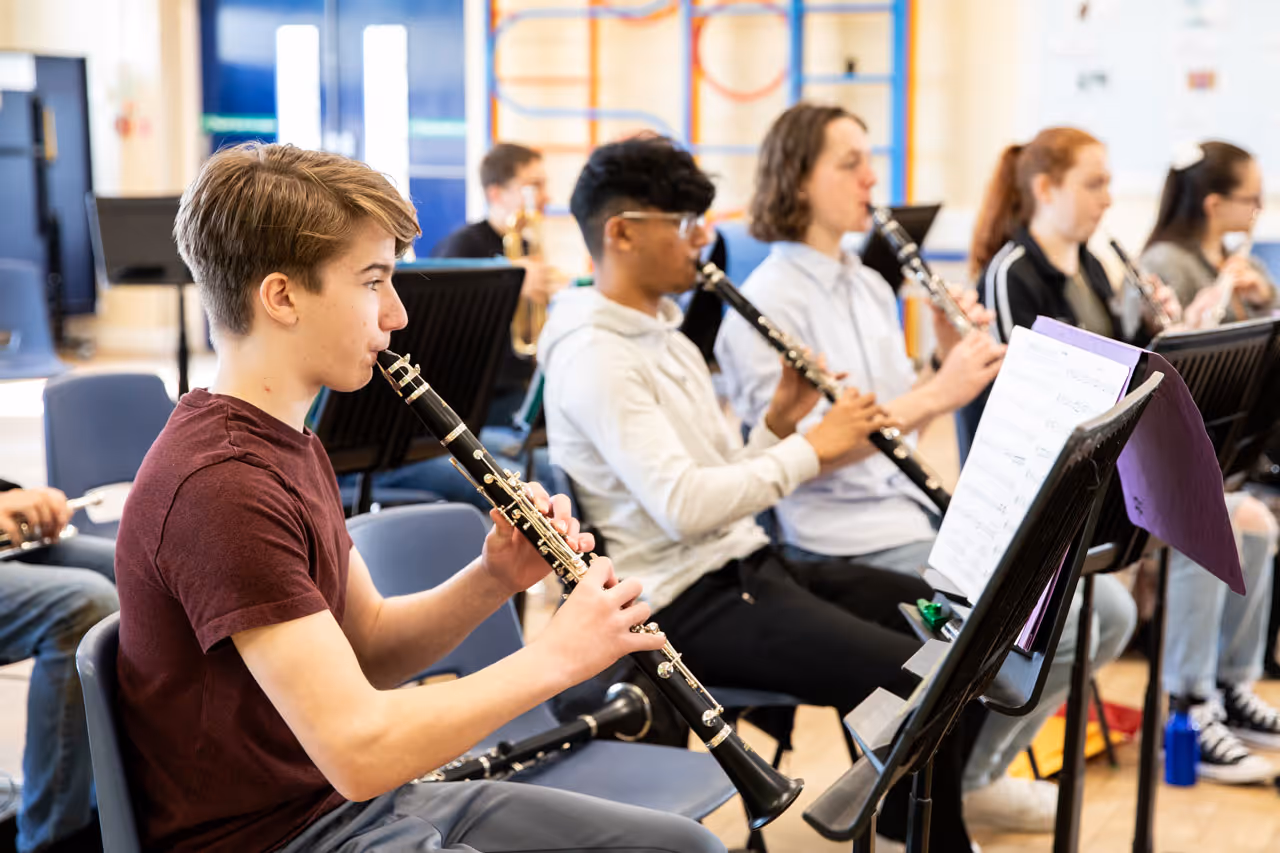 A group of young people sitting on chairs playing clarinets with sheet music on stands in front of them in a music classroom.