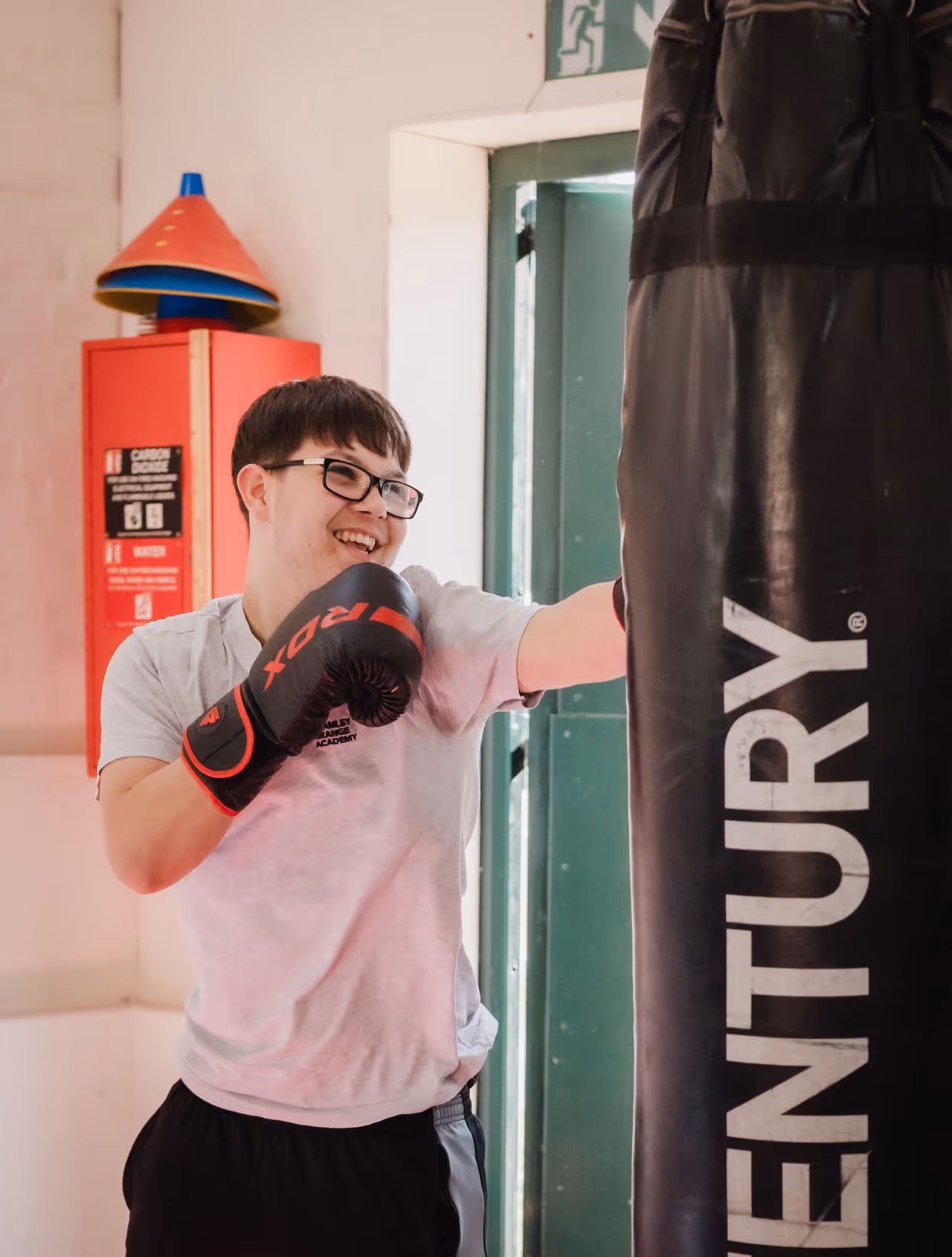 Young person wearing glasses and boxing gloves smiling while punching a Century punching bag indoors.